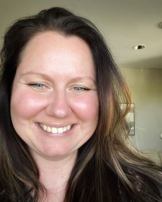 Ashleigh Pank, a midlife, cis female with she/her pronouns with long brown hair, blue eyes, Caucasian fair skin and wearing casual clothing. She is seated in a light-filled office with plants and calming decor, conveying a warm, supportive presence.