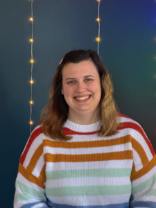 A white woman with medium-length dark hair is wearing a rainbow-striped sweater. She stands in front of a blue wall with string lights.