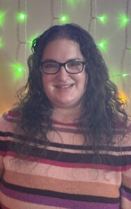 A white female with long curly hair and glasses. She is wearing a striped sweater and smiling in front of colorful string lights.