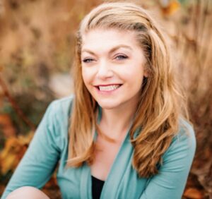 A white female with long blonde hair and blue eyes. She is smiling with her teeth showing. She is wearing a black shirt with an aqua open long sleeved shirt over it.
