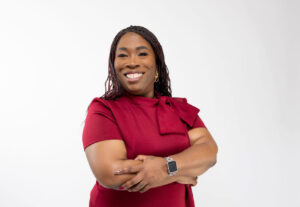 A black woman with dark brown eyes, a big smile, and long braids. She is wearing a burgundy dress with her arms folded and her background is white.