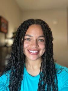 Biracial woman, young adult, green eyes and long black Locs. Wearing a blue shirt and is sitting in an office.