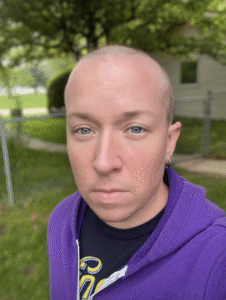 A white man with blue eyes and a shaved head. He is wearing a purple hoodie and standing outside in front of a tree.