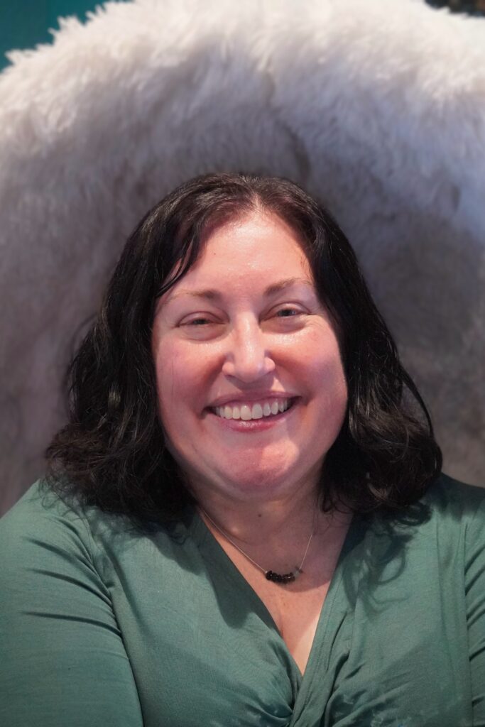 A white woman with medium-length dark brown hair wearing a green short smiling warmly.