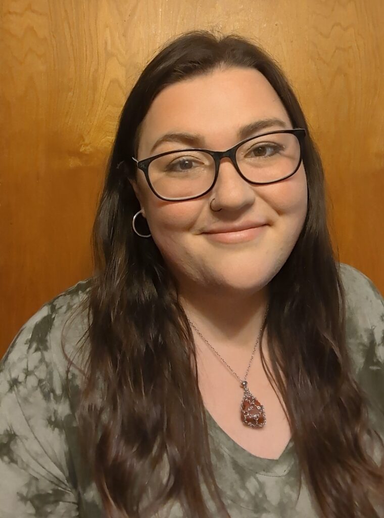 A white cisgender brunette woman with brown eyes wearing a nose piercing and glasses. She is wearing a green tie dye t-shirt and is indoors against a wood grain background.