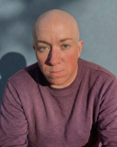 A white transgender man with a shaved head wearing an eggplant long sleeved shirt.