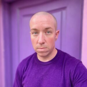 A white transgender man with a shaved head wearing a purple shirt.