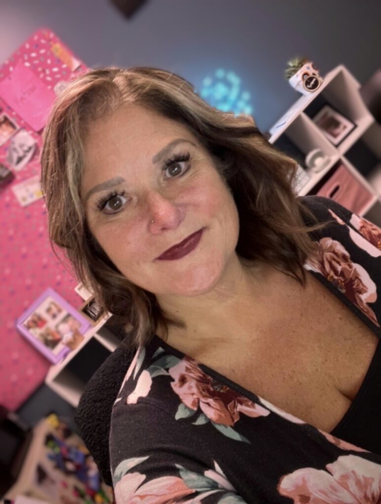 A white middle aged woman with shoulder length, wavy hair.  She is sitting in her vibrant office.