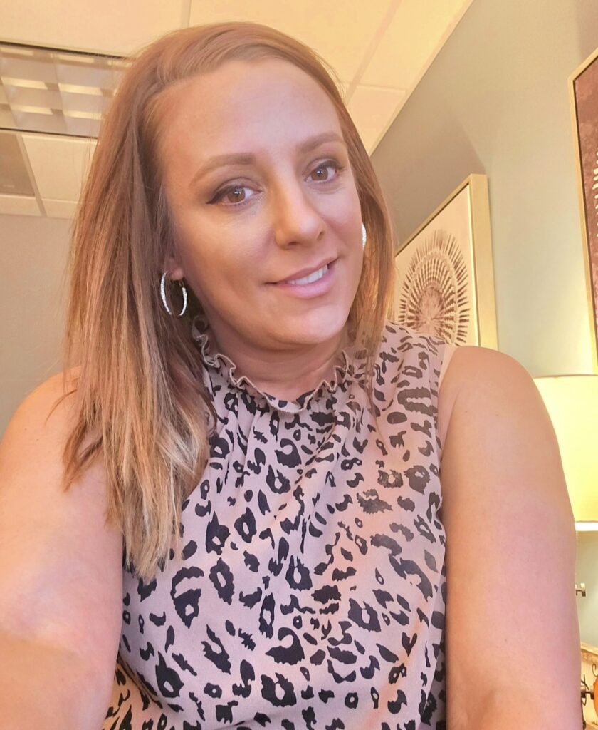 A white woman with reddish blonde hair wearing a leopard print sleeveless shirt, smiling.