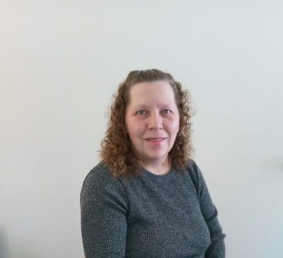 A mid life woman, smiling in front of a plain light-colored wall. She is wearing a textured dark gray top and has light brown curly hair pulled back from her face.