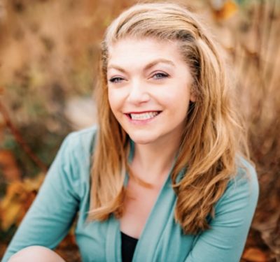 A white female with long blonde hair and blue eyes. She is smiling with her teeth showing. She is wearing a black shirt with an aqua open long sleeved shirt over it.