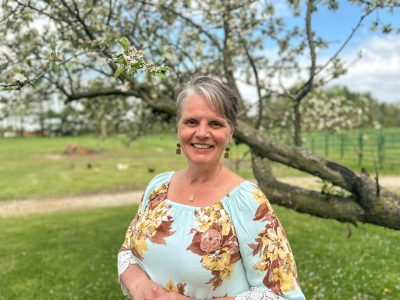 A white cisgender female with blue eyes and a short pixie haircut. She is wearing a teal floral shirt and is standing outside under an apple tree.
