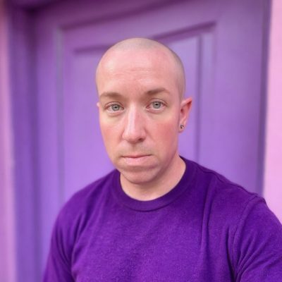 A white transgender man with a shaved head wearing a purple shirt.