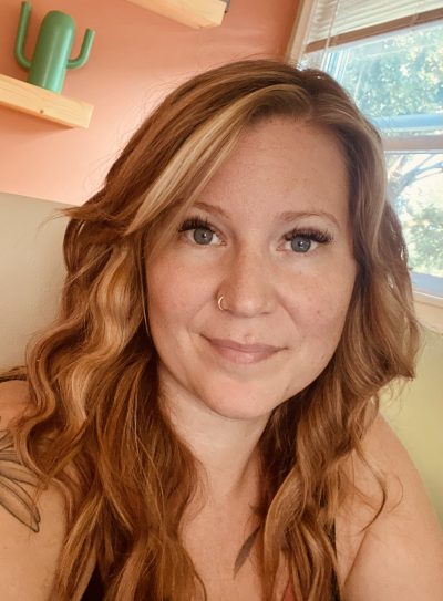 A white, midlife female with long red/blonde hair and blue eyes. She has some visible tattoos of nature and a nose piercing. She is wearing a sleeveless blouse and sitting in an office space.