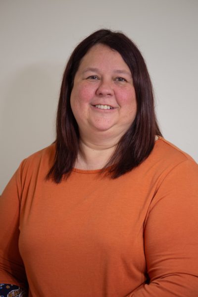A mid life woman, smiling in front of a neutral background. She is wearing a burnt orange long-sleeve top and has straight, shoulder-length dark hair.